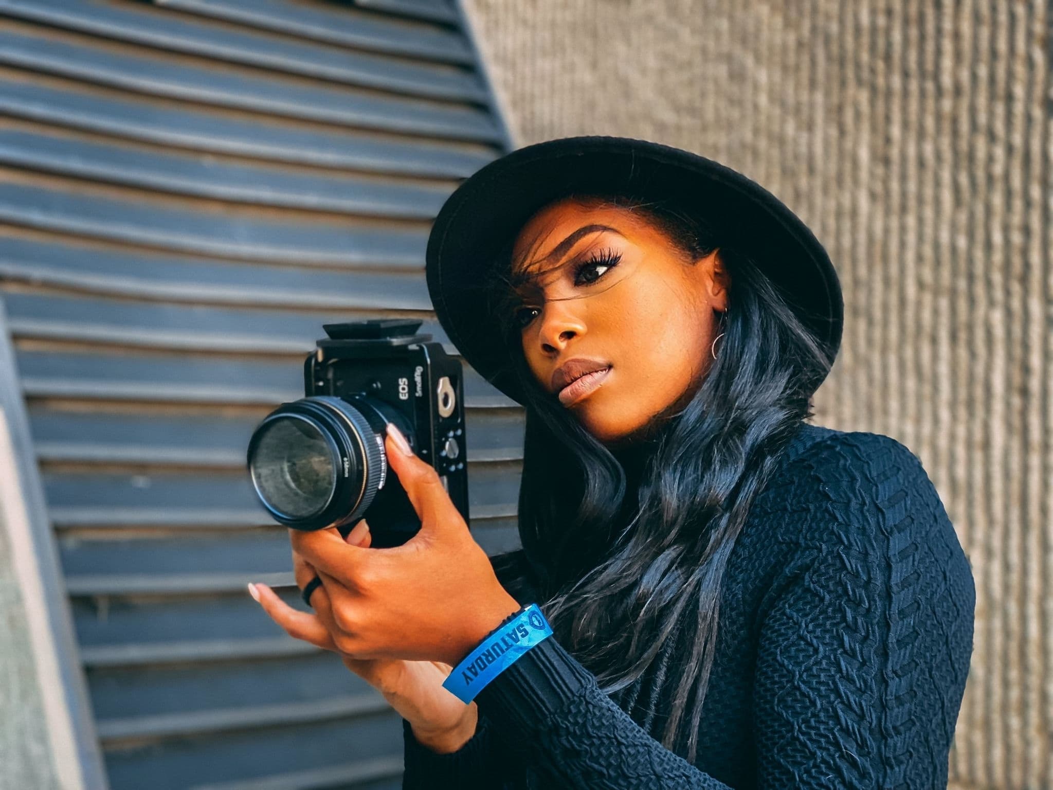 Stylish woman in a black hat and sweater holding a camera against a textured wall.