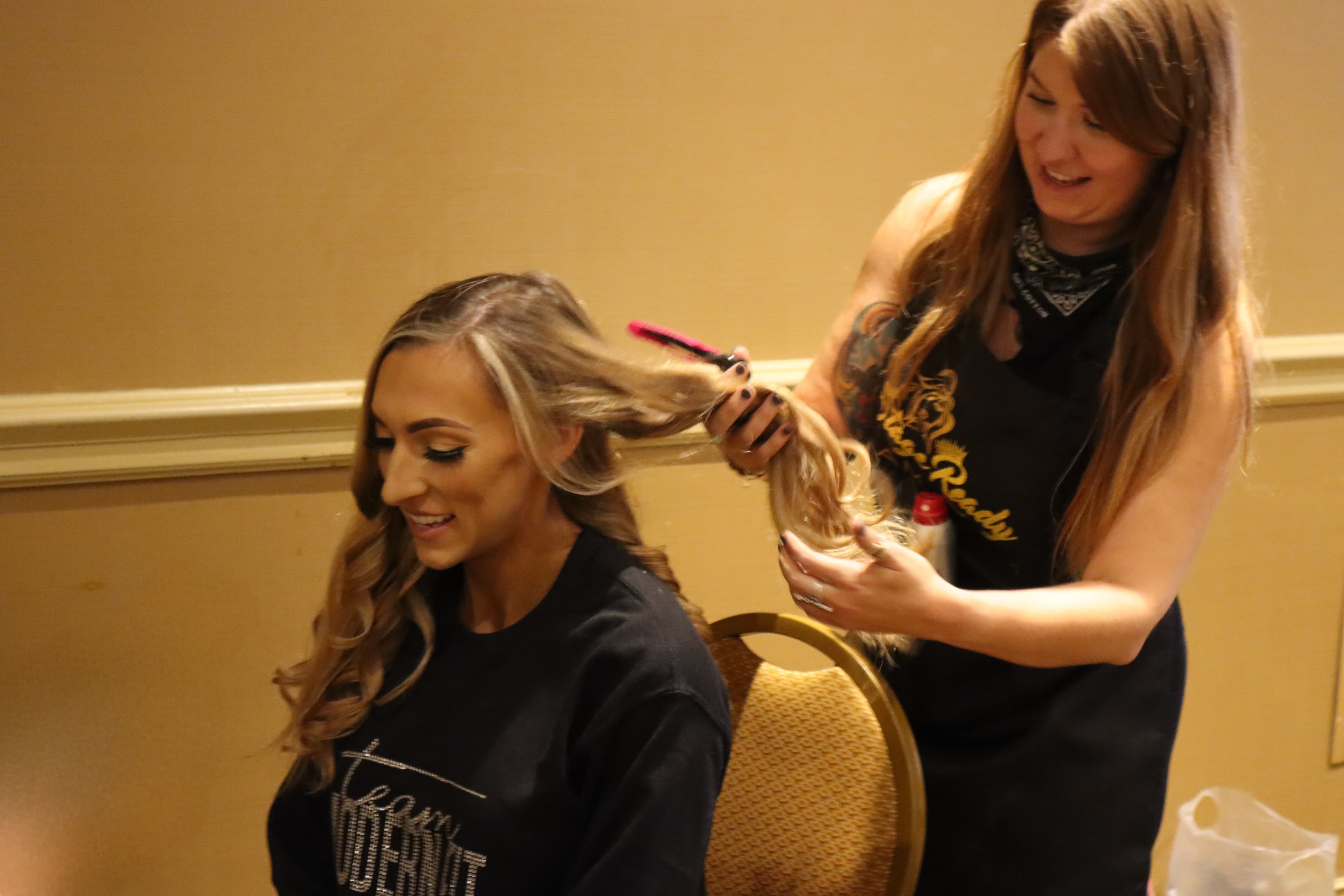 Smiling hairstylist in a black apron styles a woman's long blonde hair with a brush.