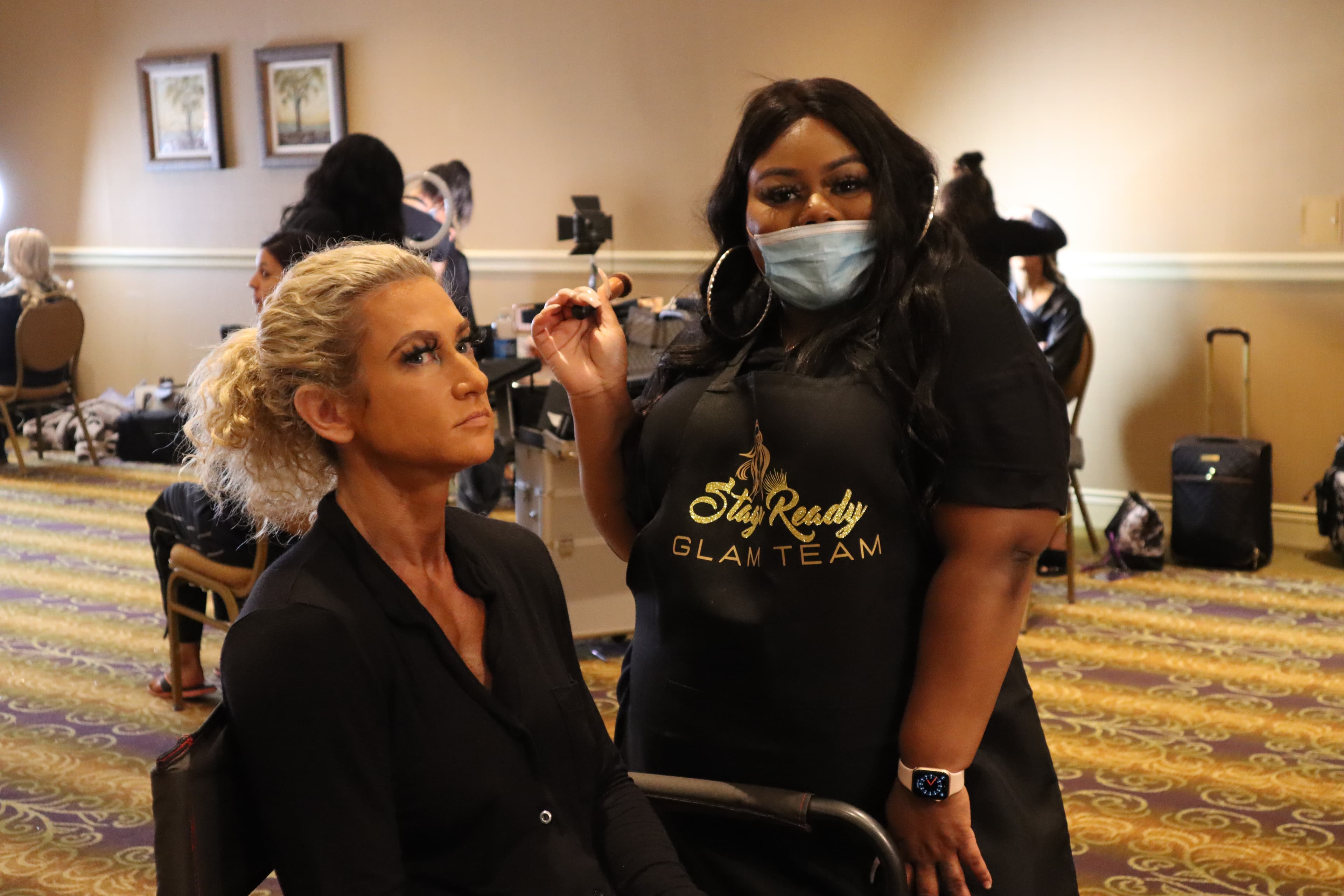 Masked makeup artist in a black apron holds a brush beside a seated blonde client.