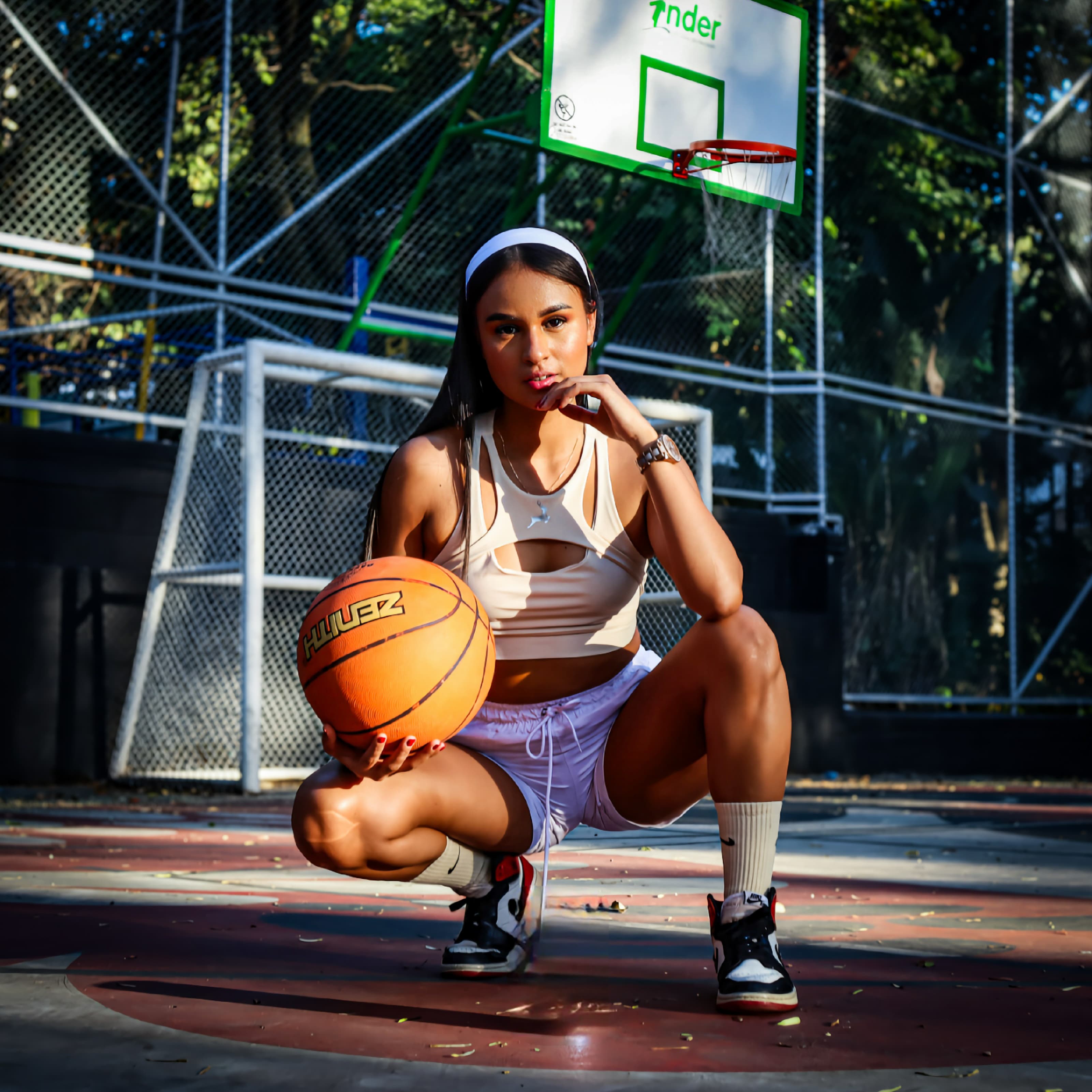 Woman in a white headband and crop top squats on a court holding a basketball.