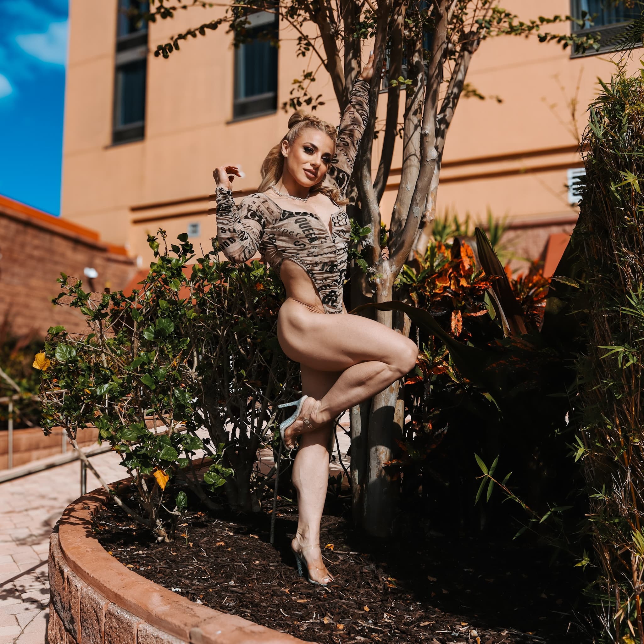 Muscular blonde woman in high-cut patterned bodysuit posing outdoors by a tree in sunlight.