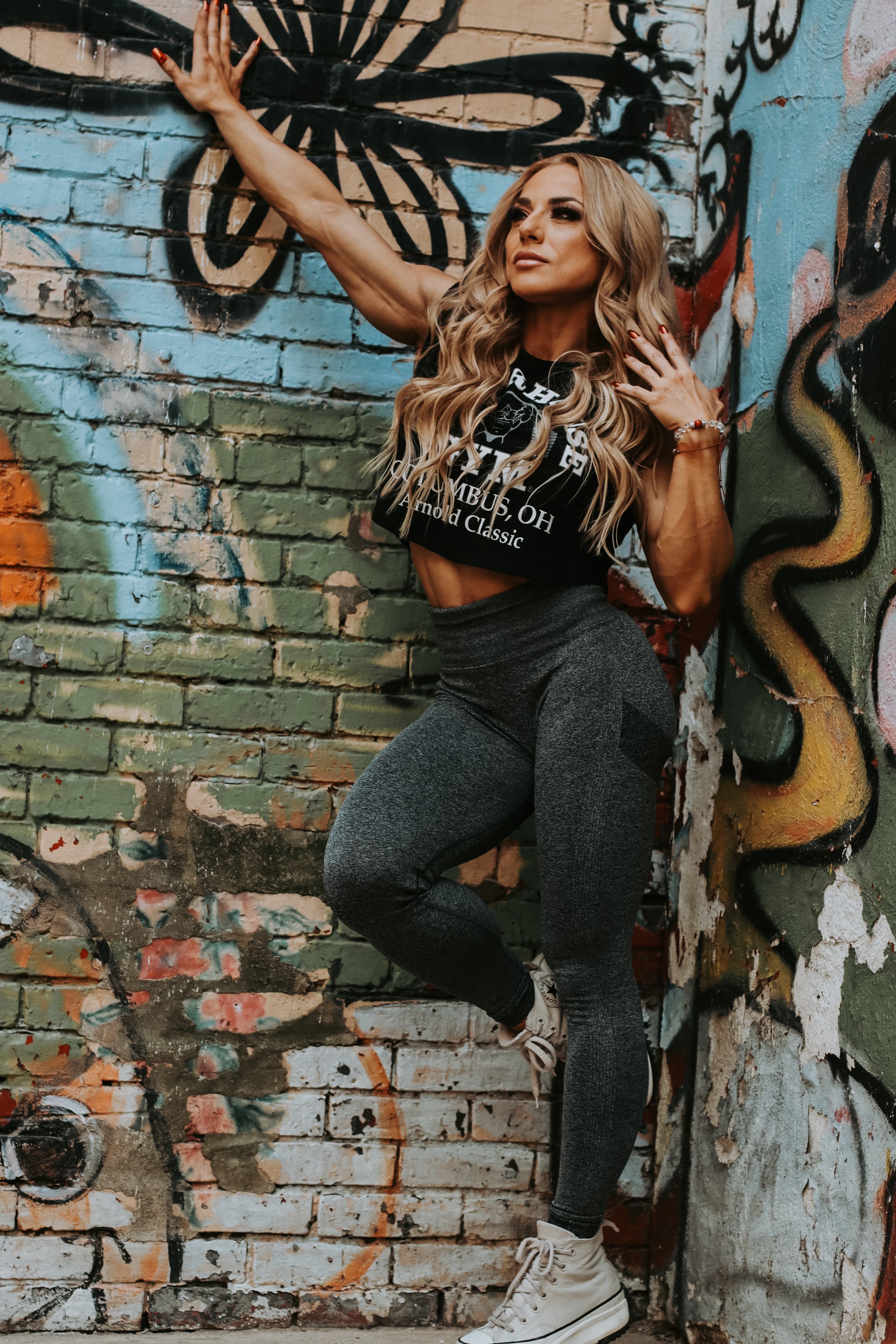 Muscular blonde woman in a black crop top posing against a colorful graffiti wall.