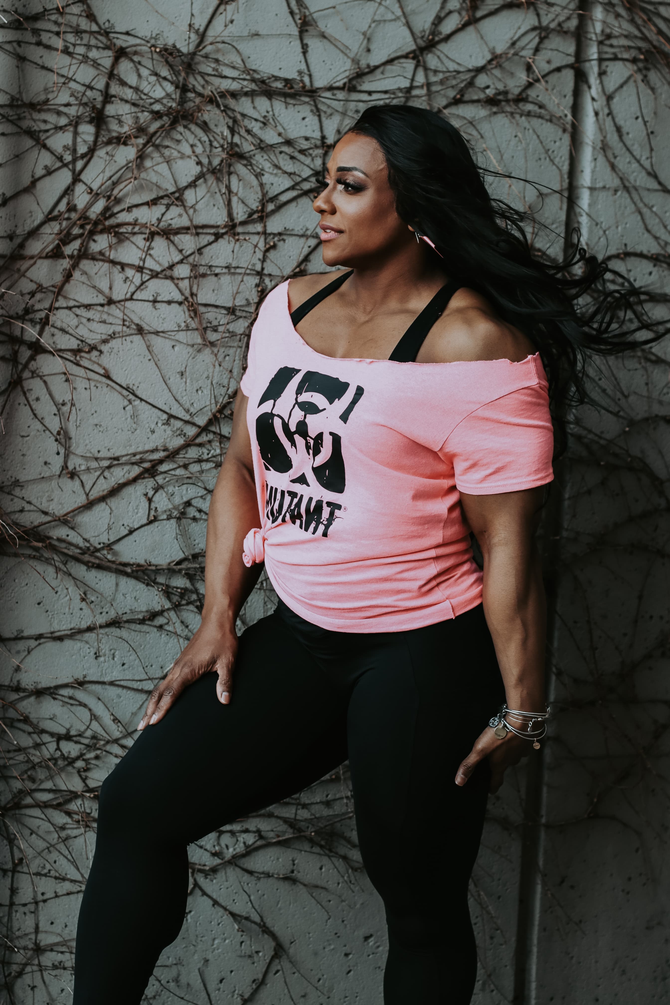 Muscular woman with long dark hair in a pink graphic tee against a vine-covered wall.