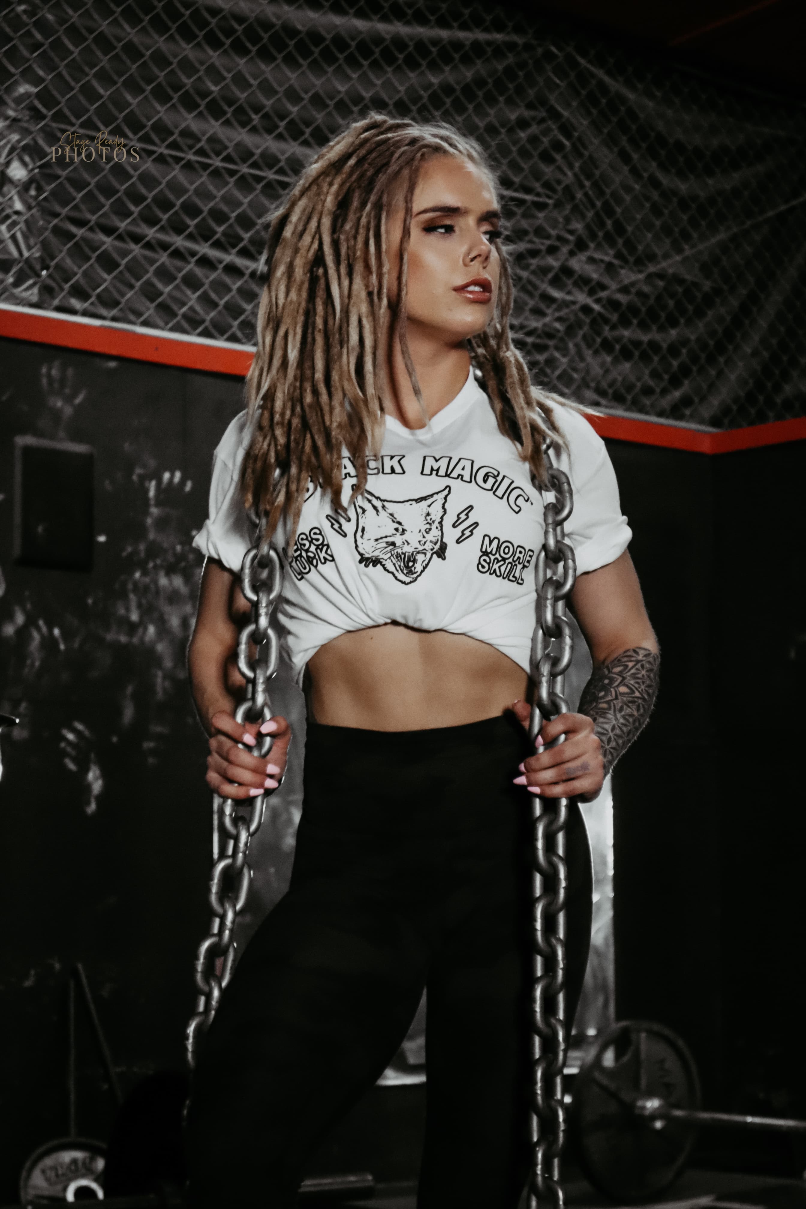 Athletic woman with blonde dreadlocks holds heavy chains in a gym, wearing a white crop top.