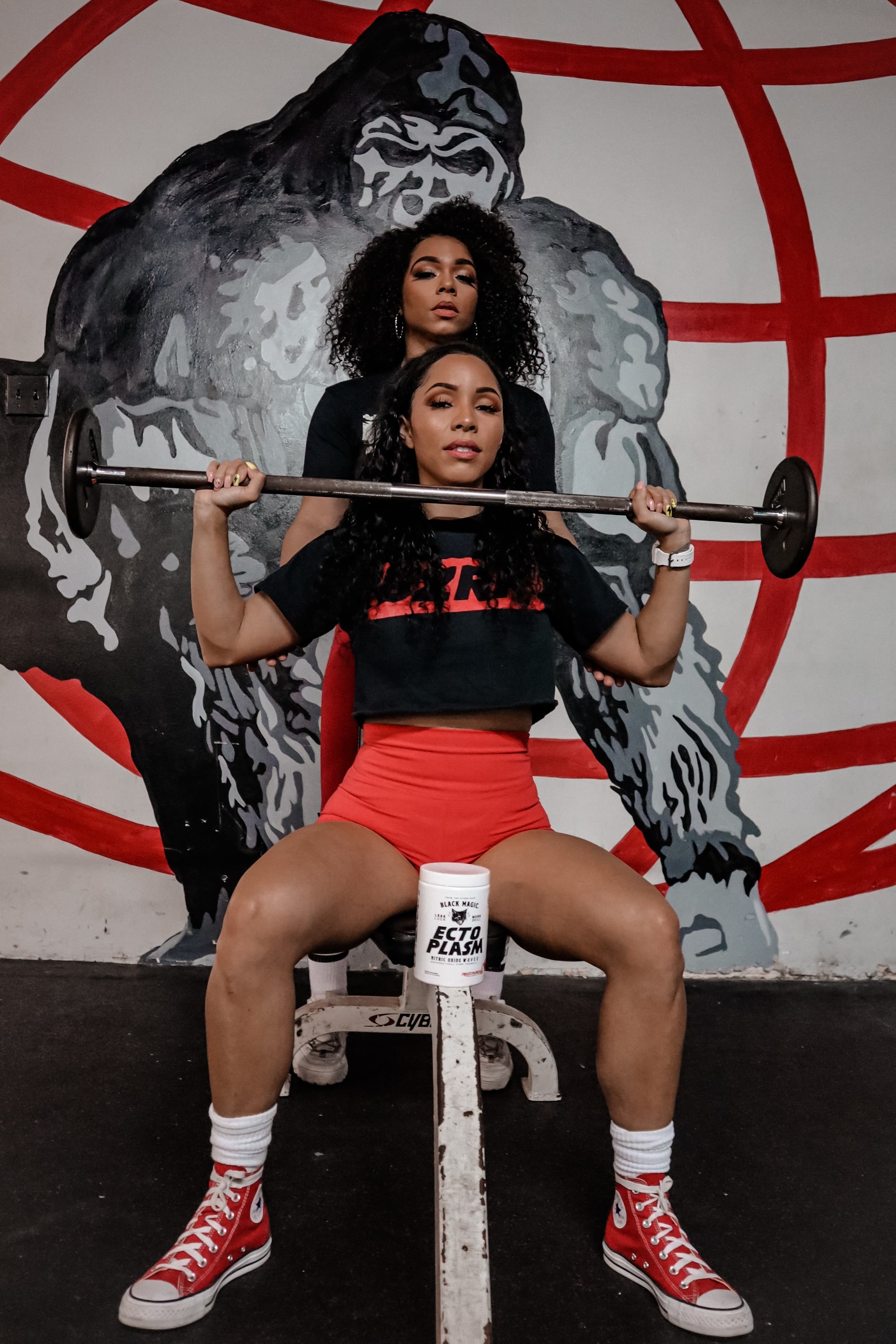 Two women pose with a barbell in front of a large, stylized gorilla wall mural.